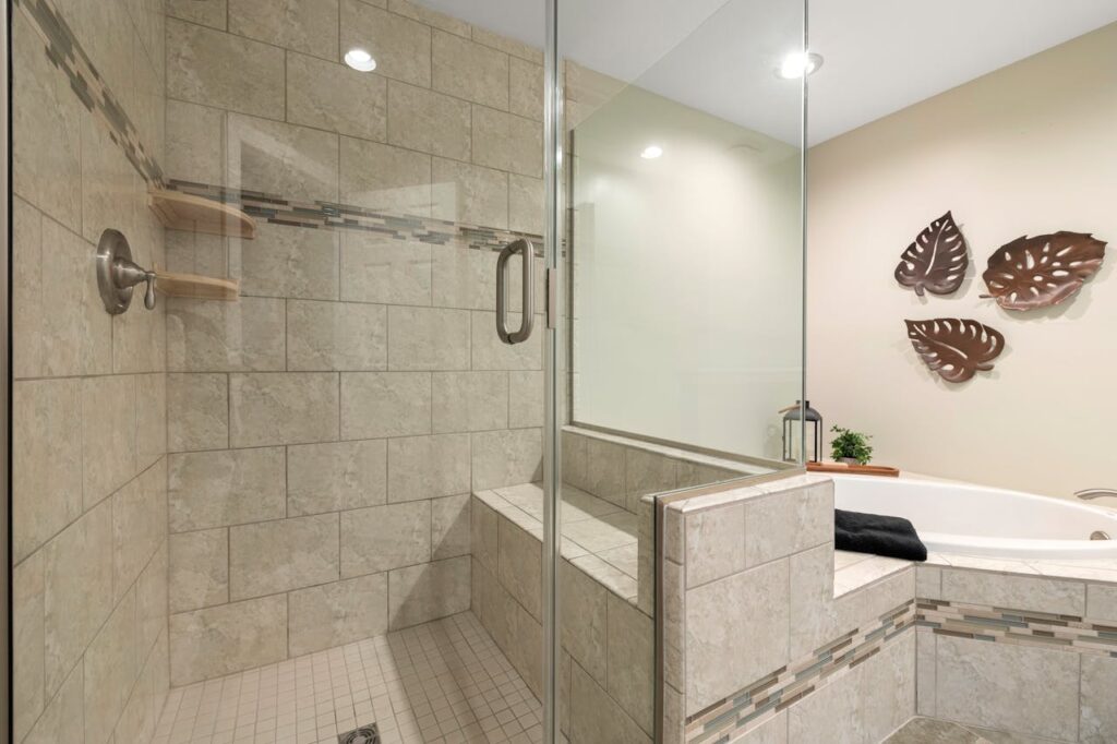 A stylish beige tiled bathroom featuring a glass shower and bathtub with decorative brown leaf art on the wall.